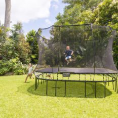 A child is jumping on a SpringFree Jumbo Square Smart Trampoline in a backyard.