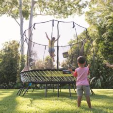 Two children playing on a SpringFree Large Oval Smart Trampoline in a backyard.