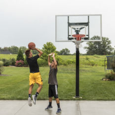 Two boys playing ALL-AMERICAN in a backyard with a basketball hoop.