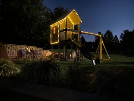 A VinylNation Royal Cottage Clubhouse with lights in the yard at night.